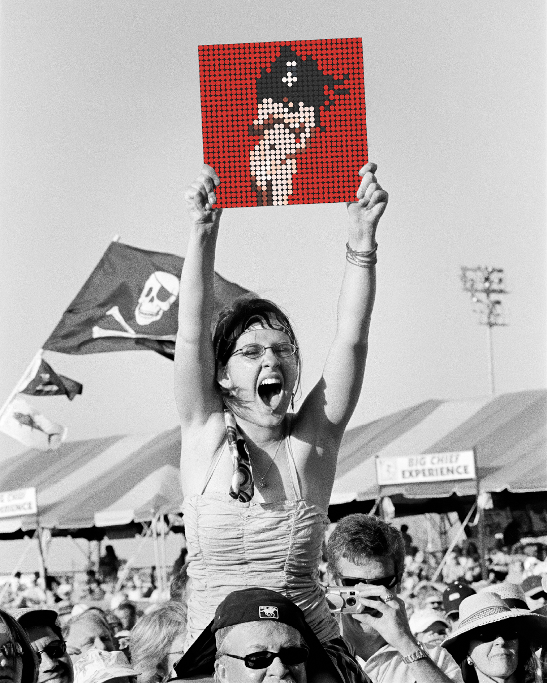 Fotografía en blanco y negro de una multitud en un festival. En el centro, una joven emocionada se encuentra sobre los hombros de otra persona, levantando con entusiasmo un cartel a color que muestra un mosaico de piezas estilo LEGO con la silueta de la portada del álbum MOTOMAMI + de Rosalía. Al fondo se ven carpas, banderas ondeando (incluyendo una de calavera pirata) y personas tomando fotos. La imagen celebra el fanatismo y la energía de los conciertos.