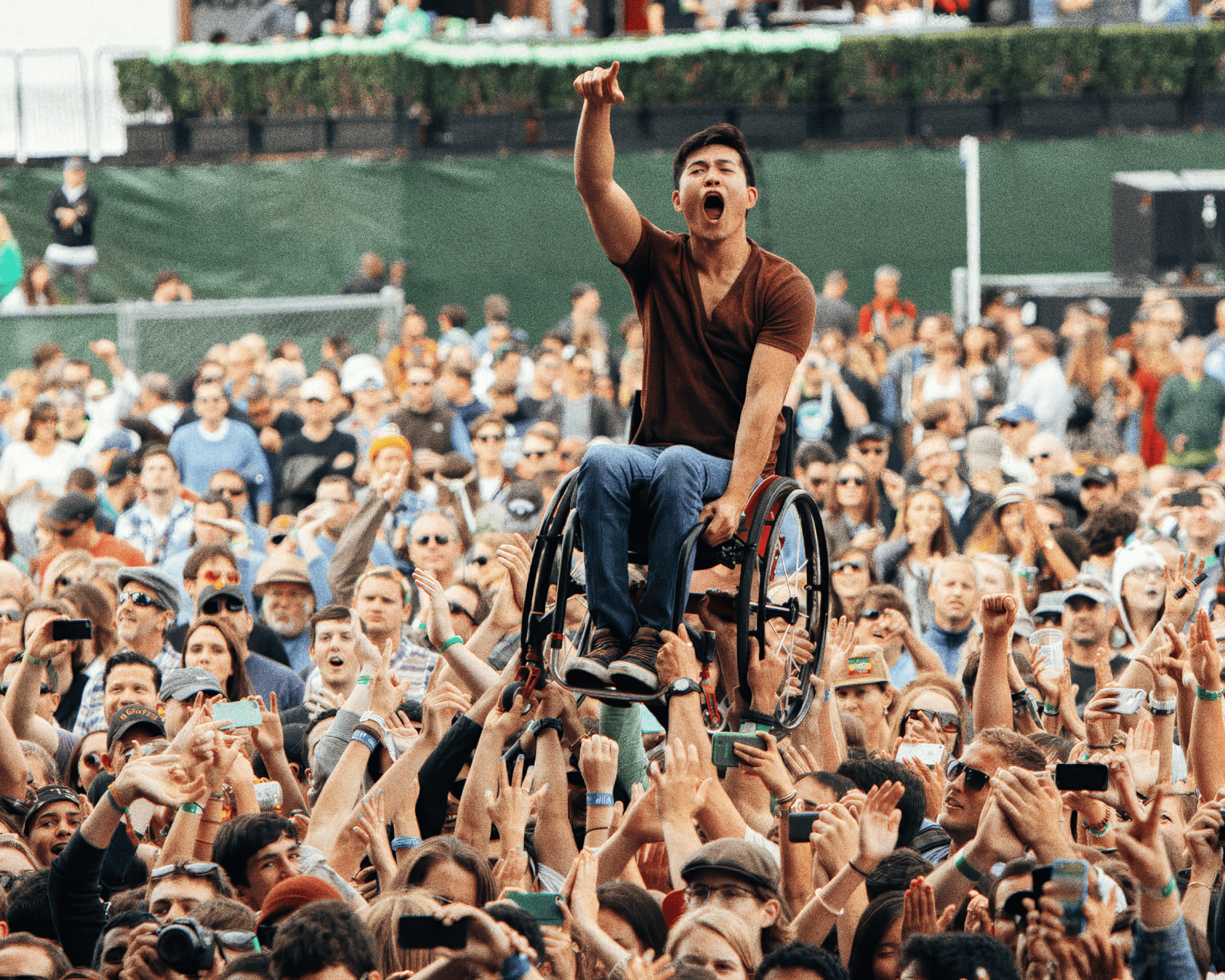 Un hombre en silla de ruedas es elevado por la multitud durante un concierto o festival. El hombre, con una camiseta burdeos y pantalones vaqueros, señala al frente mientras grita con entusiasmo. La gente a su alrededor sonríe, levanta los brazos y captura el momento con sus teléfonos móviles. La imagen transmite un ambiente de inclusión, euforia y conexión colectiva.